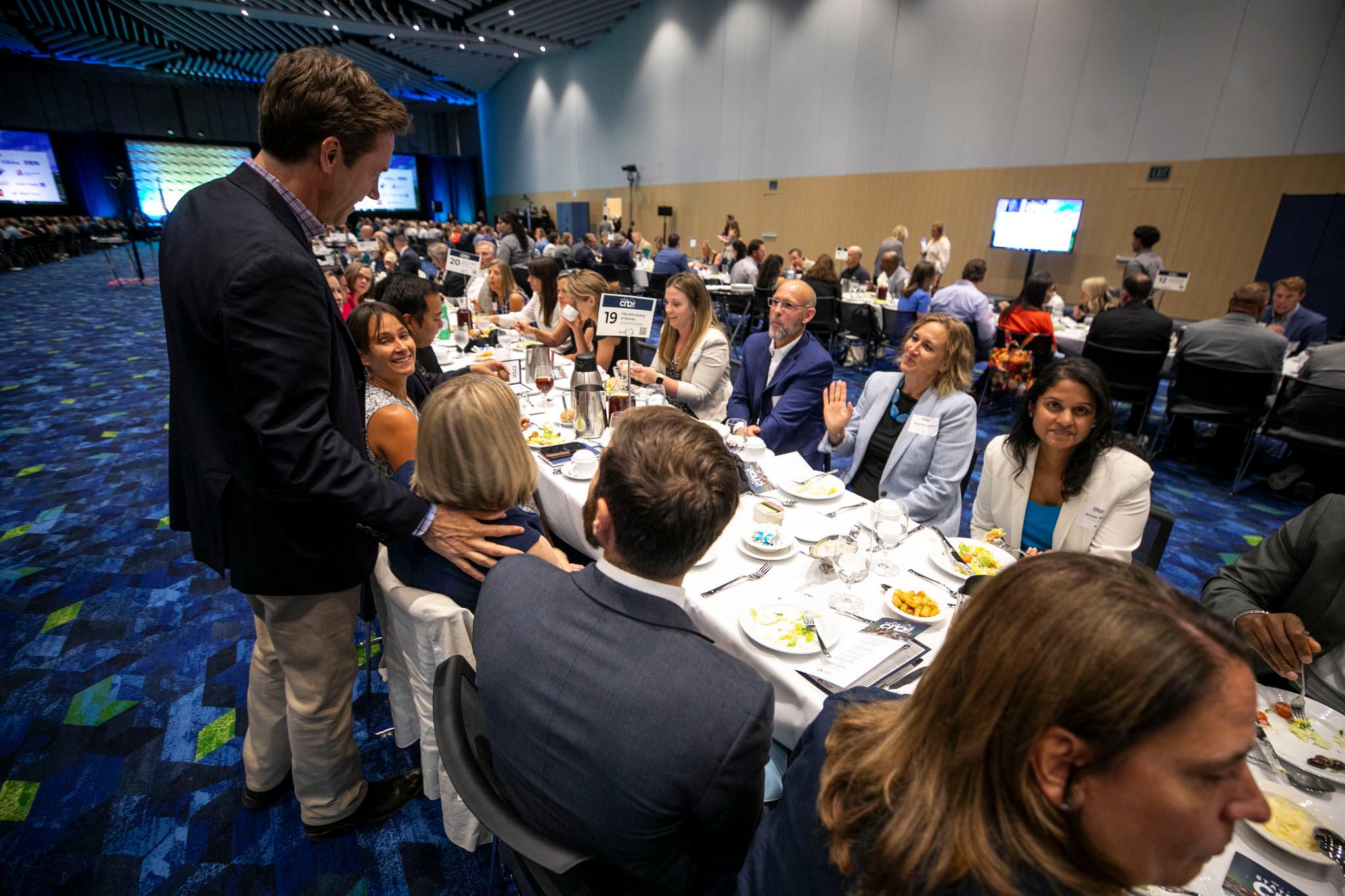 A man in a blazer stands over a table of other well-dressed people eating lunch; one person waves at him.