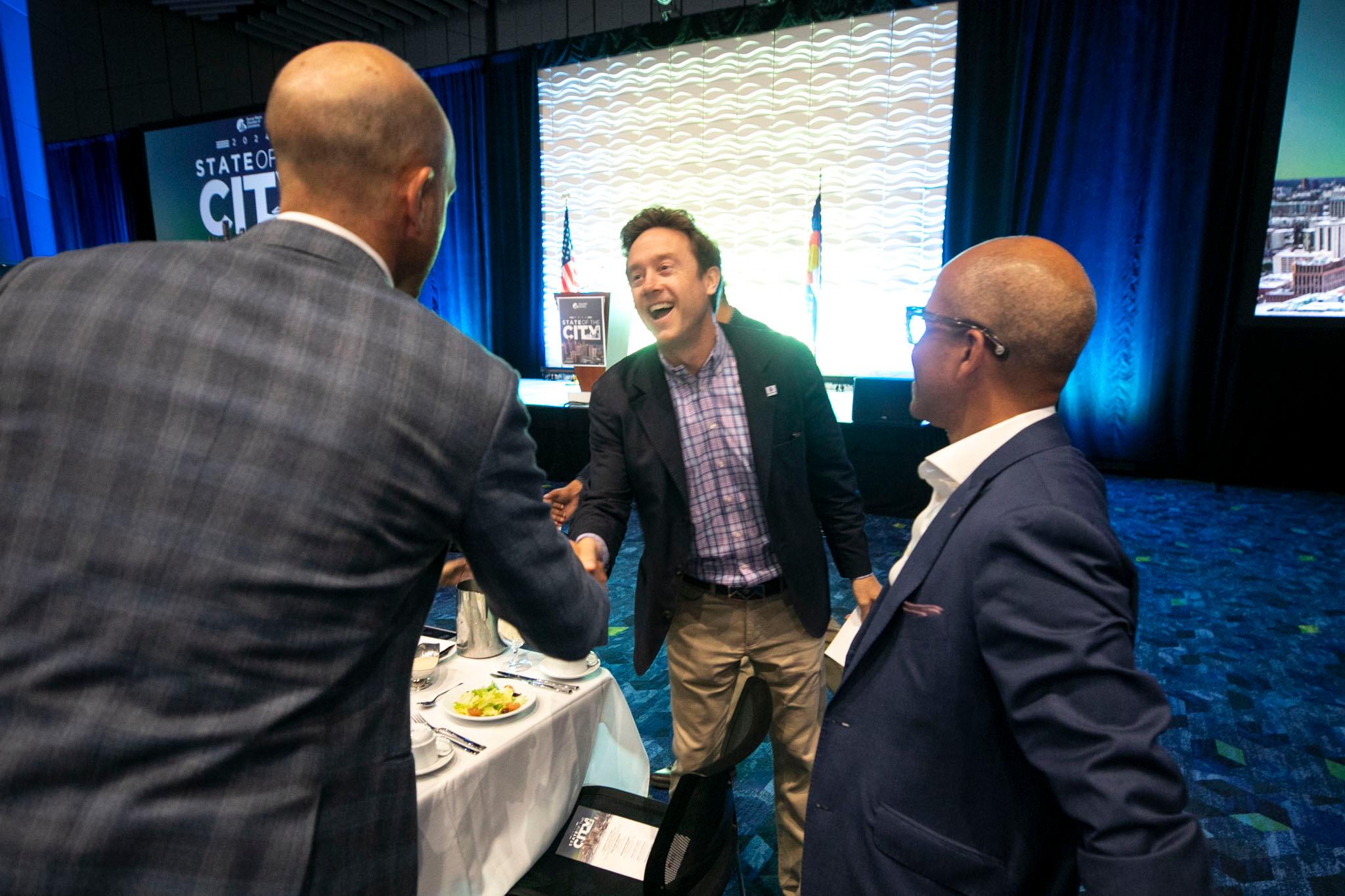 A man in a sportscoat laughs as he greets two bald men in suits.