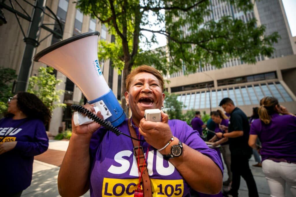 A woman in a purple SEIU shirt smiles widely as she yells into a megaphone.