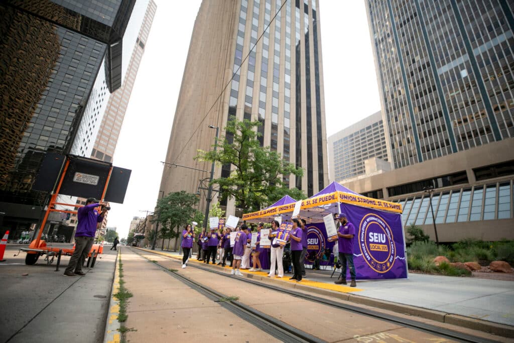 A group of people in purple shirts stand in front of purple tents on a city sidewalk.