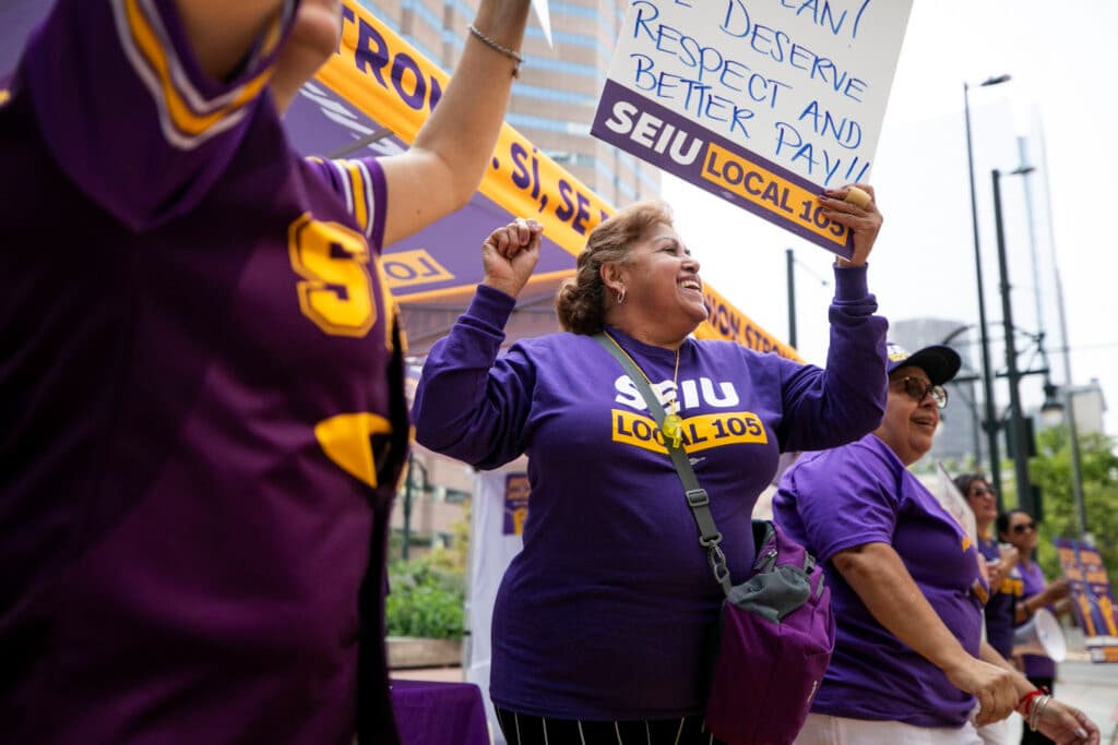 A woman in a purple SEIU shirt smiles as she raises one fist and holds a protest over her head with her other hand. She's surrounded by people doing the same.