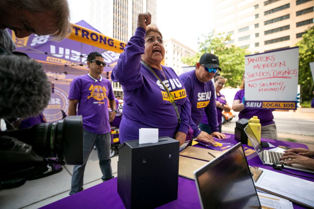 A woman in a purple SEIU shirt raises a fist as she shouts; there's a ballot box in front of her with a paper ballot sticking halfway out.