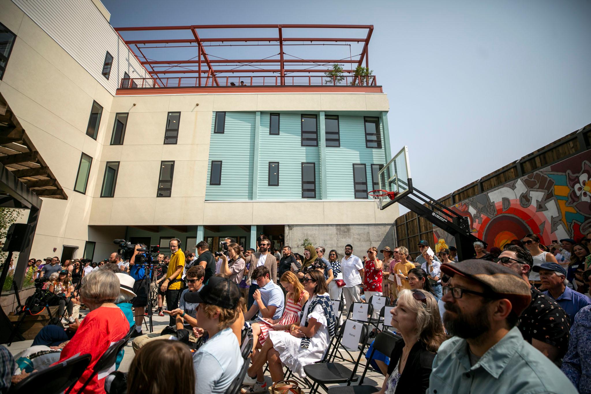 A large crown of people stands on a patio, beneath a new building colored in beige, turquoise and rust.