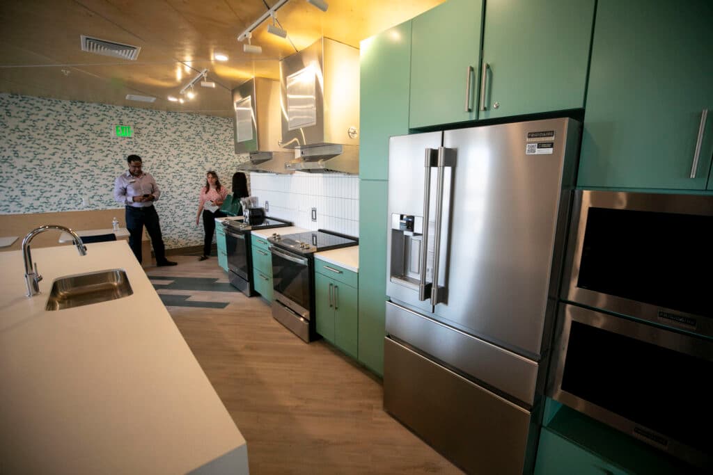 A silver refrigerator is embedded amid seafoam cabinets, next to new ovens and microwaves.