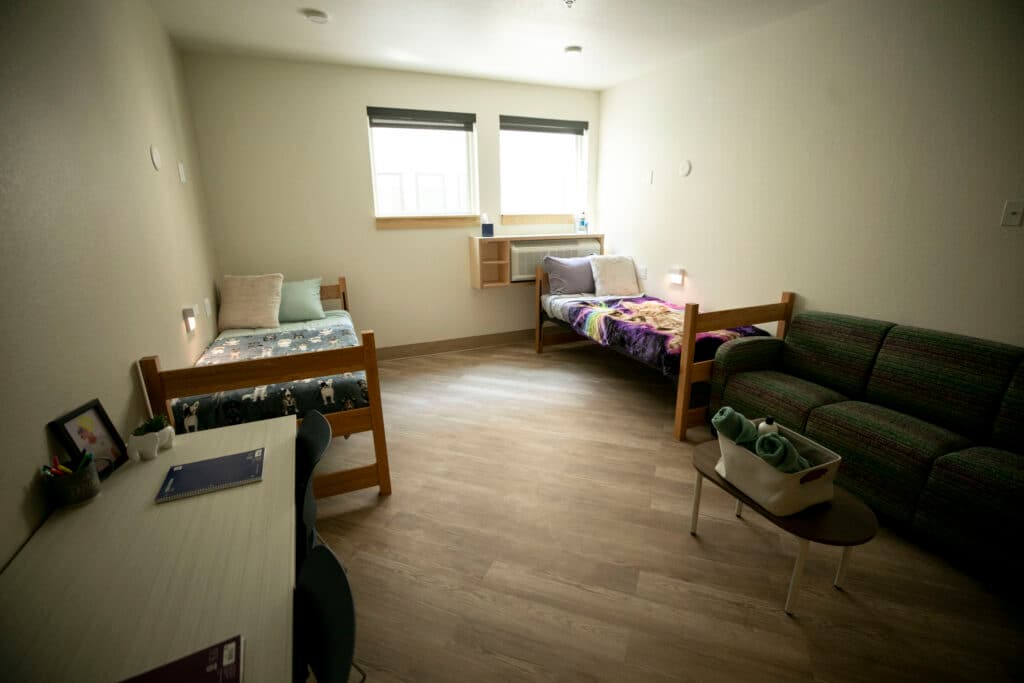 Light pours into a bedroom from a read window, illuminating two small wooden beds covered with colorful blankets, a couch, coffee table and desk.