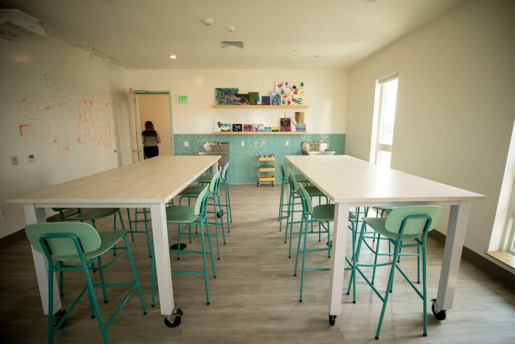 A bright room with tall craft tables surrounded by seafoam bar stools; art supplies line shelves in the back.