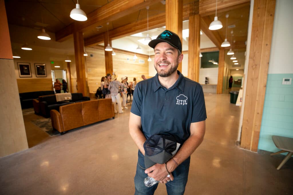 A man in a polo shirt and ballcap smiles widely inside a large, warm room lined with wood beneath tall ceilings.