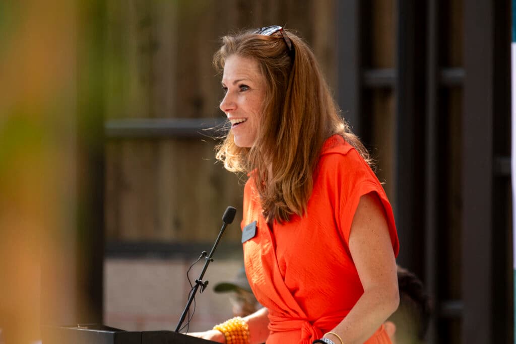 A woman in a bright red dress smiles behind a podium, gazing towards an audience out of frame.