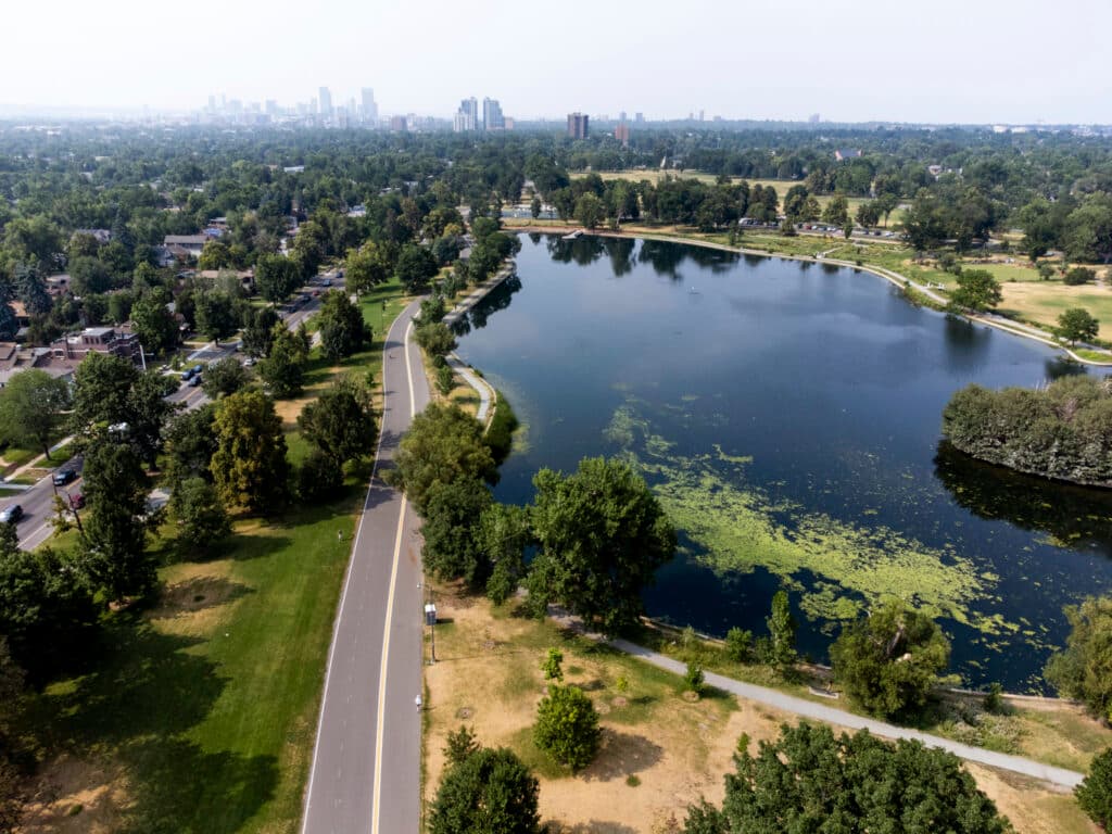 An aerial view of a big lake surrounded by trees; a road runs along its left side; a cityscape fades in the distance.