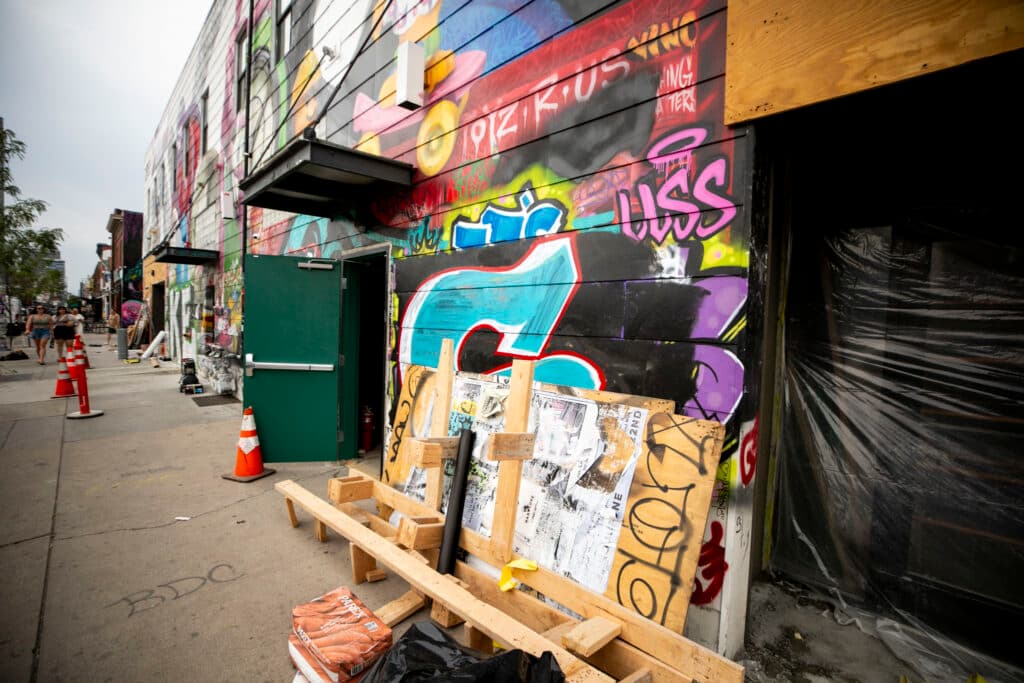 Wood is stacked against the exterior of a building, which is covered in paint — you can see that some art has been covered by newer layers of paint.