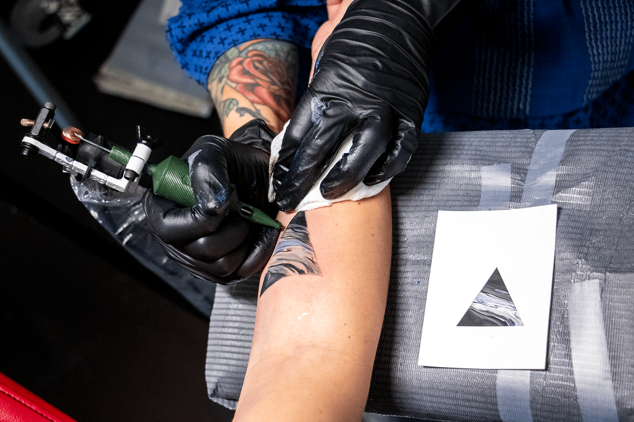 An arm lays on a table as a pair of black gloved hands tattoo right above the inner elbow a triangle - with a photograph of a triangle laying next to the arm.