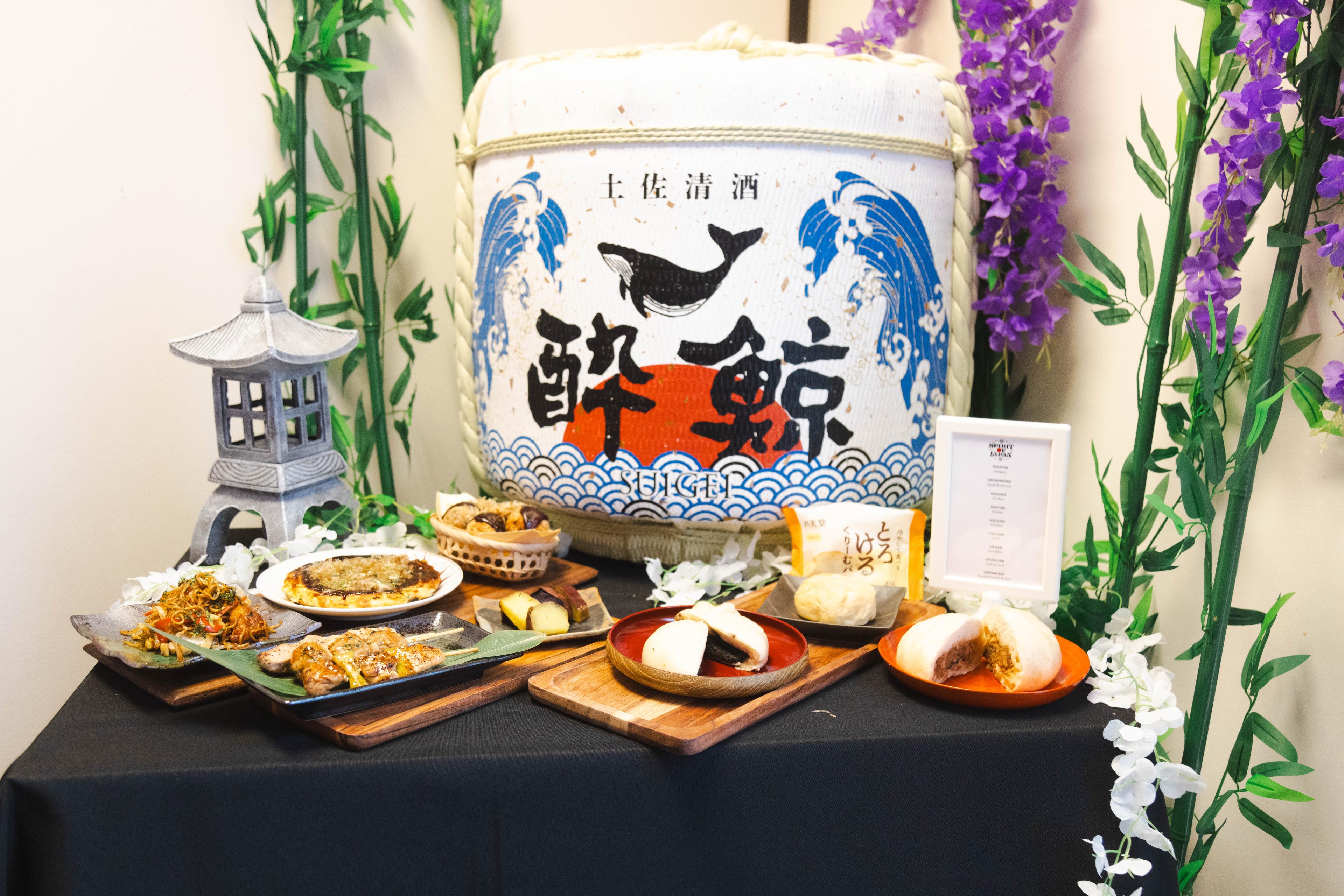 An assortment of food options that will be available at the Spirit of Japan festival, displayed on a table in front of a white ceramic pot with a humpback whale and Japanese characters painted on it. Food includes noodles, buns, yakitori and mochi.