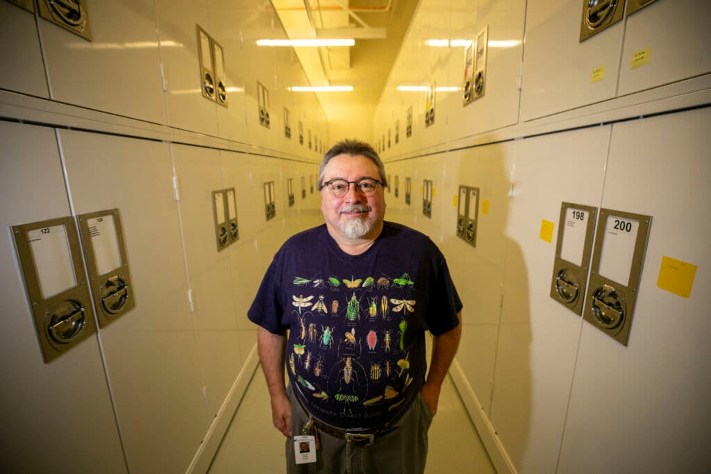 A man in a shirt filled with images of bugs stands in an aisle of white cabinets that stretches far behind him.