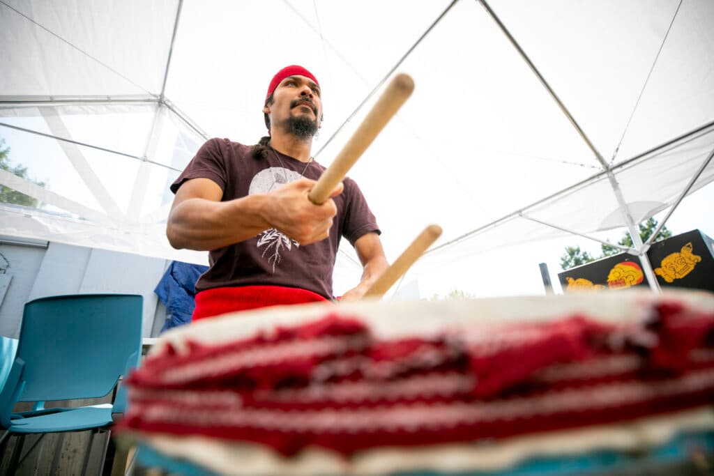 A man in a red headband stares into the middle distance as he bangs a drum with wooden sticks in his hand. The drum is out of focus in the foreground; we look up at him.