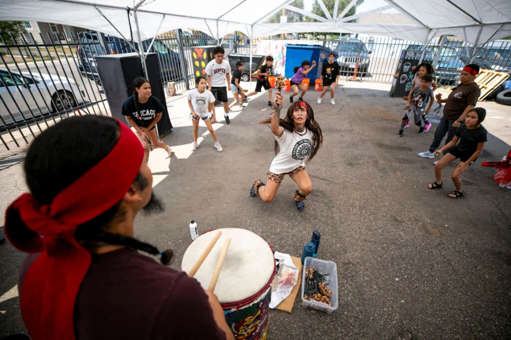 A view over the shoulder of a man in a red headband who's drumming with two wooden sticks. Beyond him, a woman looks up, crouching as she dances in a circle of kids who match her moves.