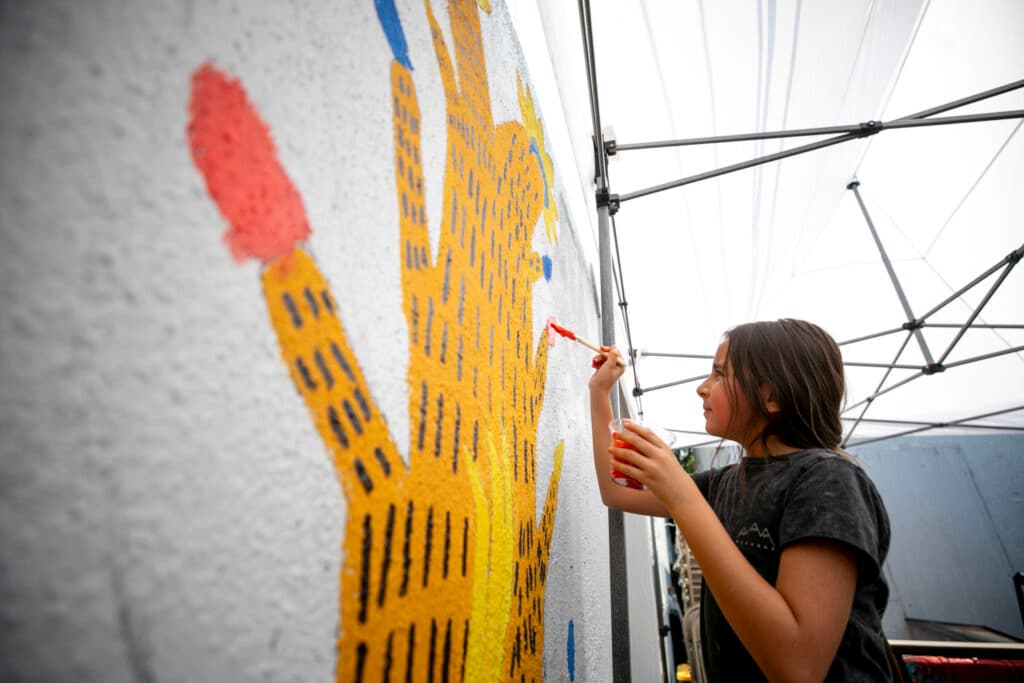 A girl looks up at a yellow mural as she positions an orange-tipped brush towards it; it appears to be the top of a husk of corn, and she's painting a flower on one of its tips.