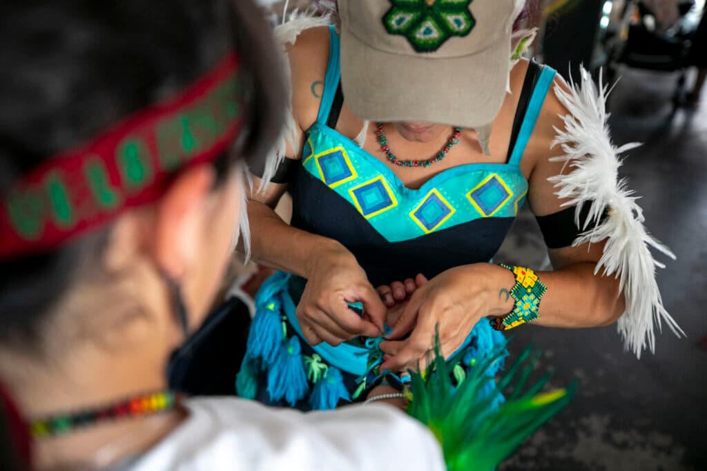 A top-down view as a woman in a colorful teal costume ties a string on the wrist of someone standing out of focus in the foreground: hands touching hands in preparation.