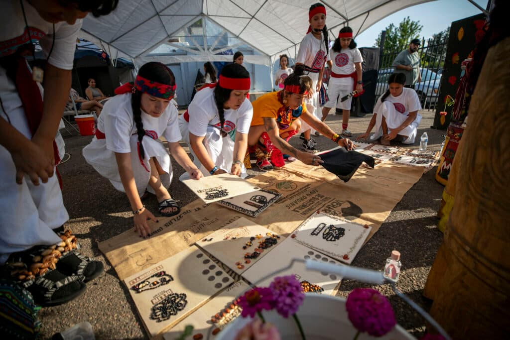 A line of people, mostly women, wearing white costumes and red headbands kneel in low evening light, adding wooden art pieces to a burlap mat. A woman in yellow in the middle spreads a black cloth over it.