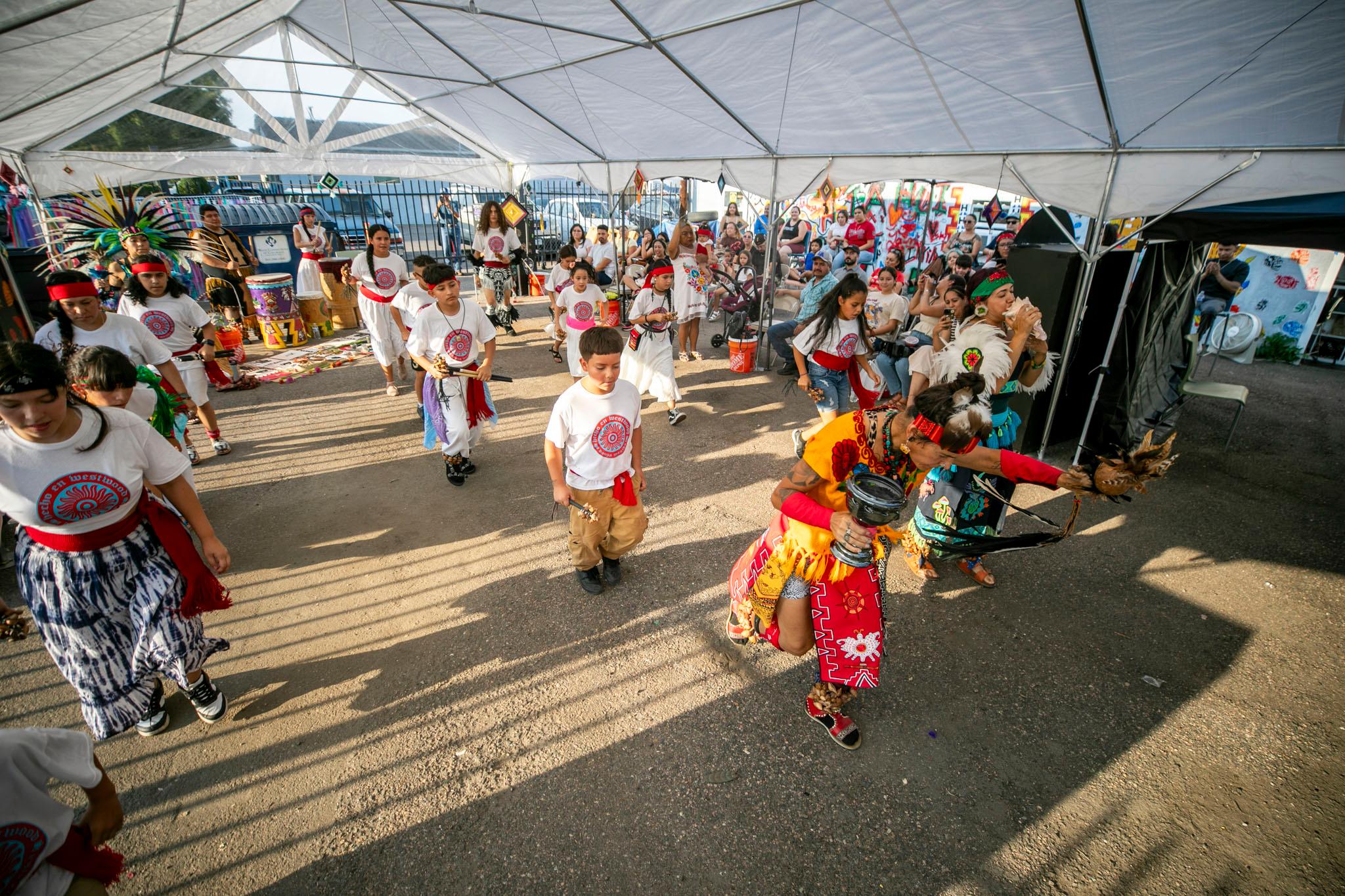 A group of kids in white are frozen in motion behind a woman in a yellow costume who's mid-lunge as she dances. They stand on asphalt, under a white tent; an audience can be seen in the background.