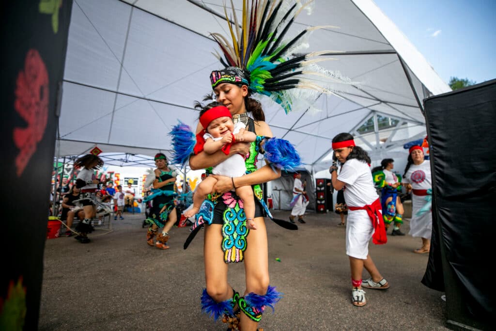 A woman in a tall, feathered headdress holds a baby wearing a red headband; they dance along with a crowd of kids behind them.