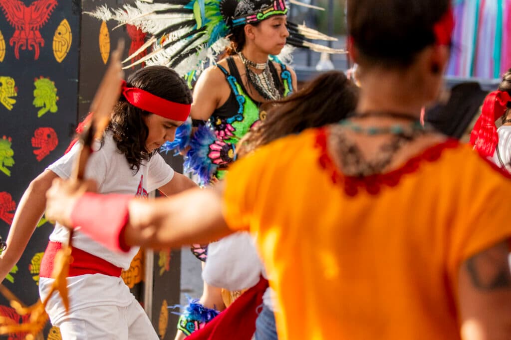 A young man in a red headband is deep in concentration as he dances, surrounded by other people in motion who wear costumes in yellow, green and teal.