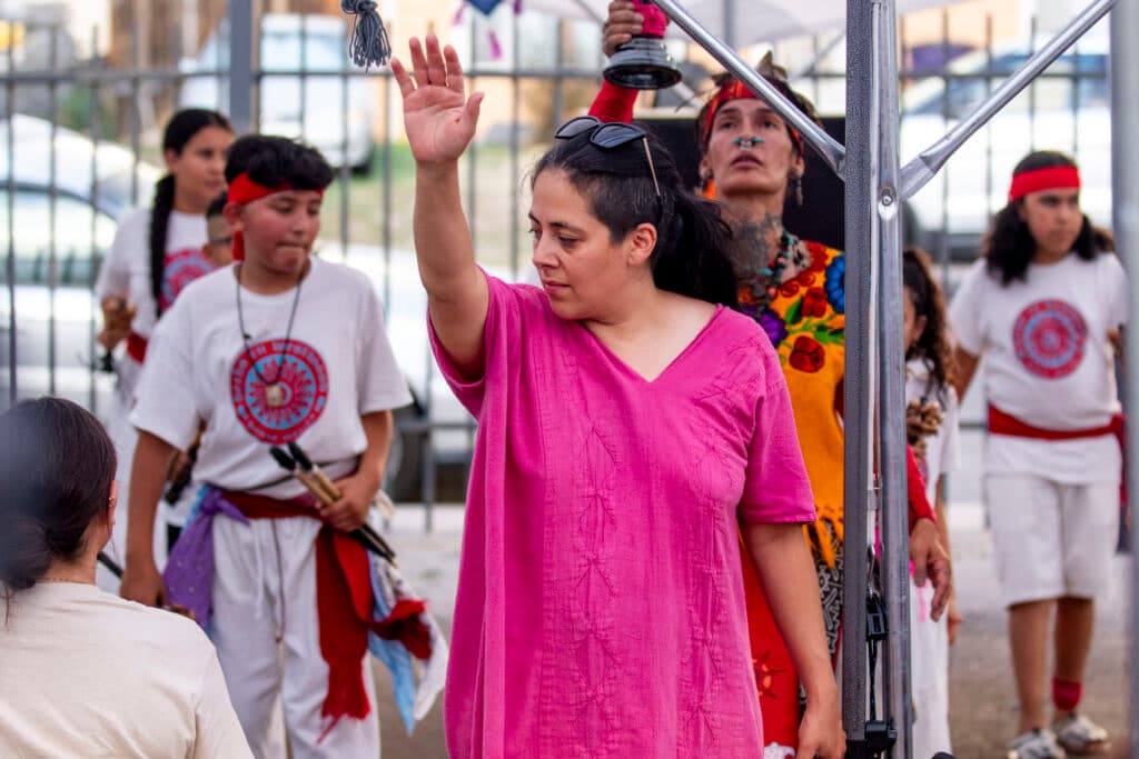 A woman in a pink dress holds up one hand; behind her are kids in white and a woman in yellow, who raises a cup to the sky.