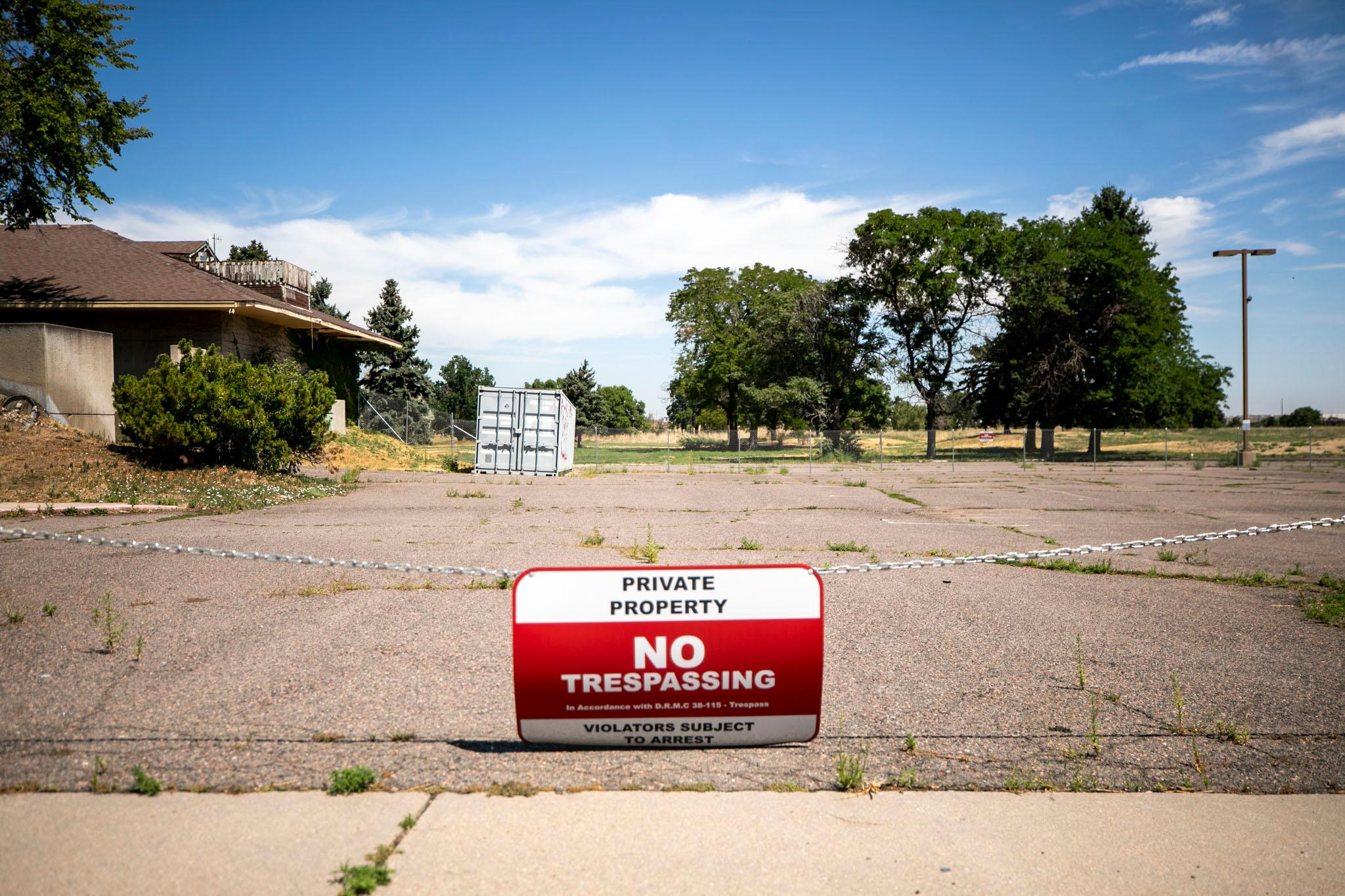 A parking lot is blocked by a chain and red and white sign in the foreground that reads "NO TRESPASSING." In the background, we can see a green and yellow lawn, trees and a building.