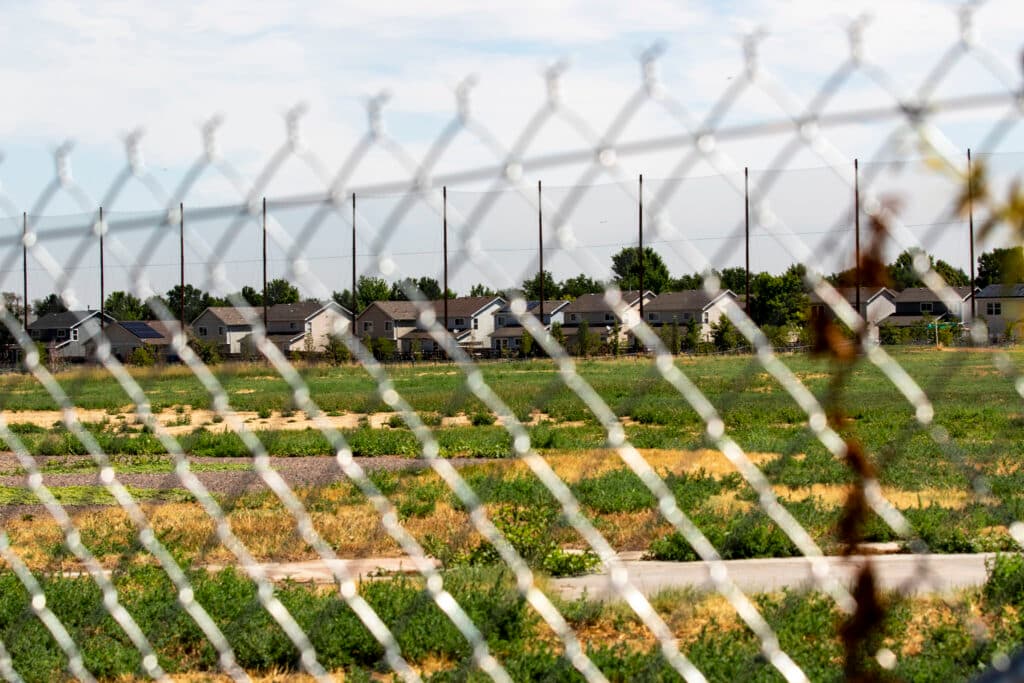 An out-of-focus fence in the foreground patterns the image of a green and yellow field with homes rising on the horizon.