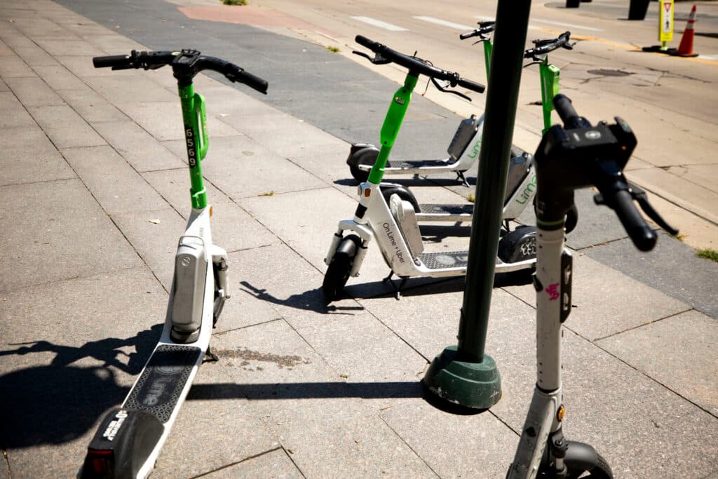 A collection of scooters, most white with green accents, on a stone tile sidewalk.