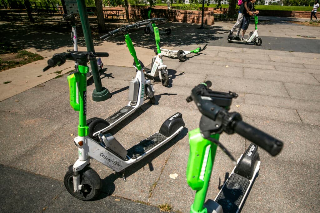 A collection of scooters, most white with green accents, on a stone tile sidewalk.