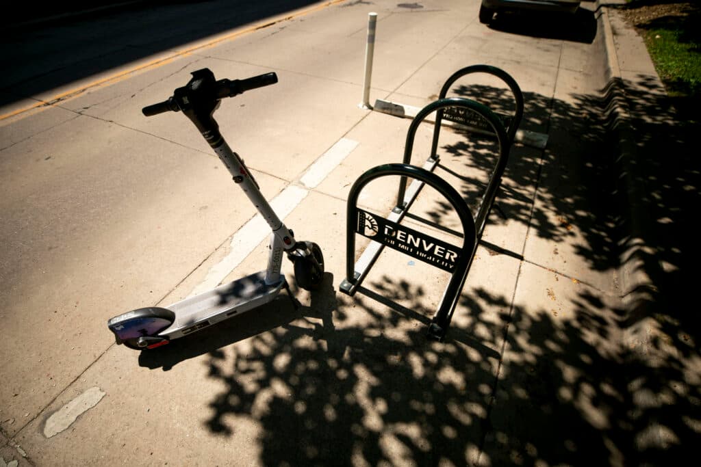 A scooter is silhouetted by a brightly lit street. Next to it is a bike rack, also silhouetted, with cut-out lettering that reads &quot;DENVER.&quot;