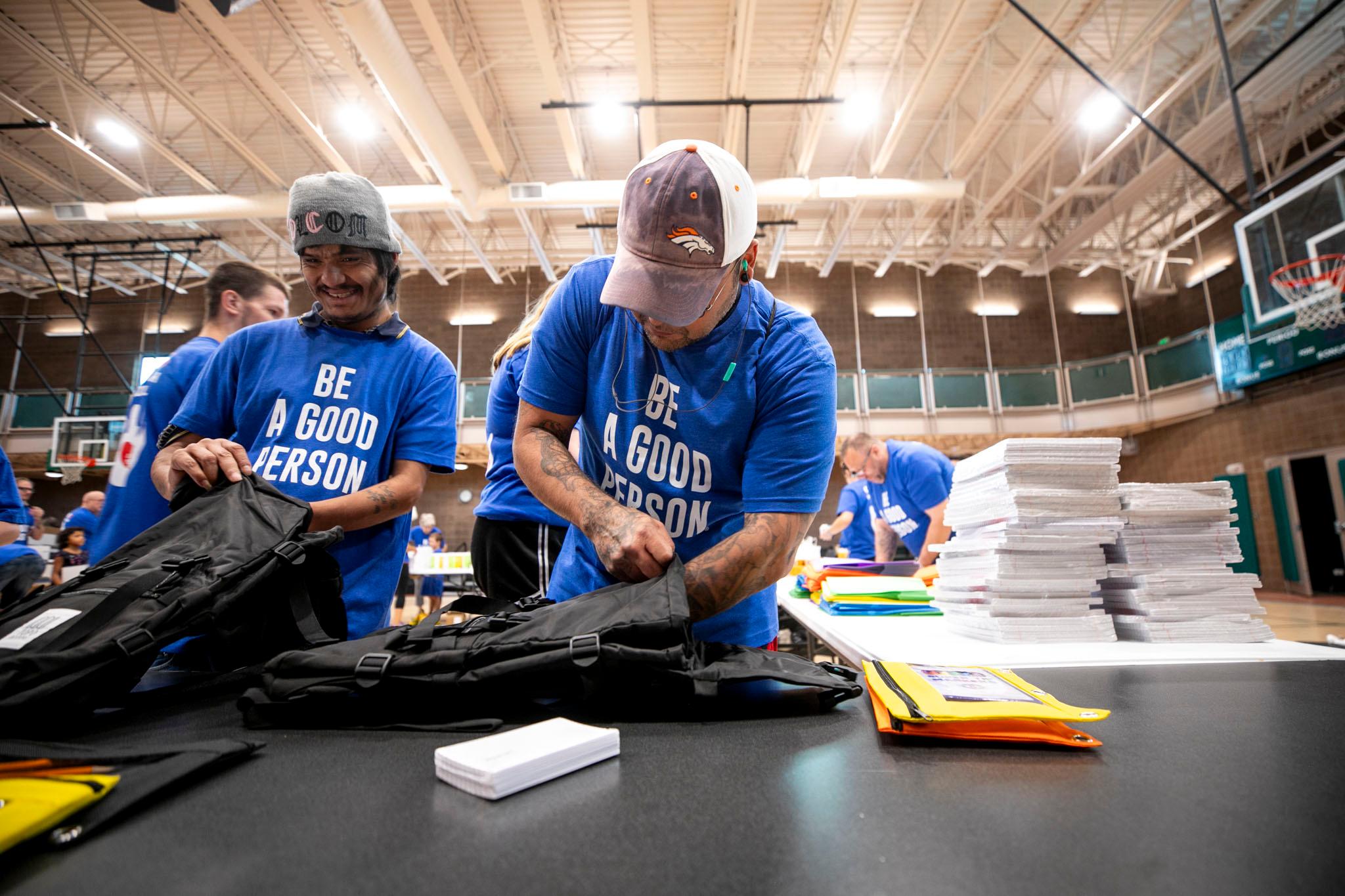 A man in a blue t-shirt that reads "BE A GOOD PERSON" sticks his hand into a black backpack. He stands in a gym and is surrounded by school supplies, stacks of paper and brightly colored pen pockets.