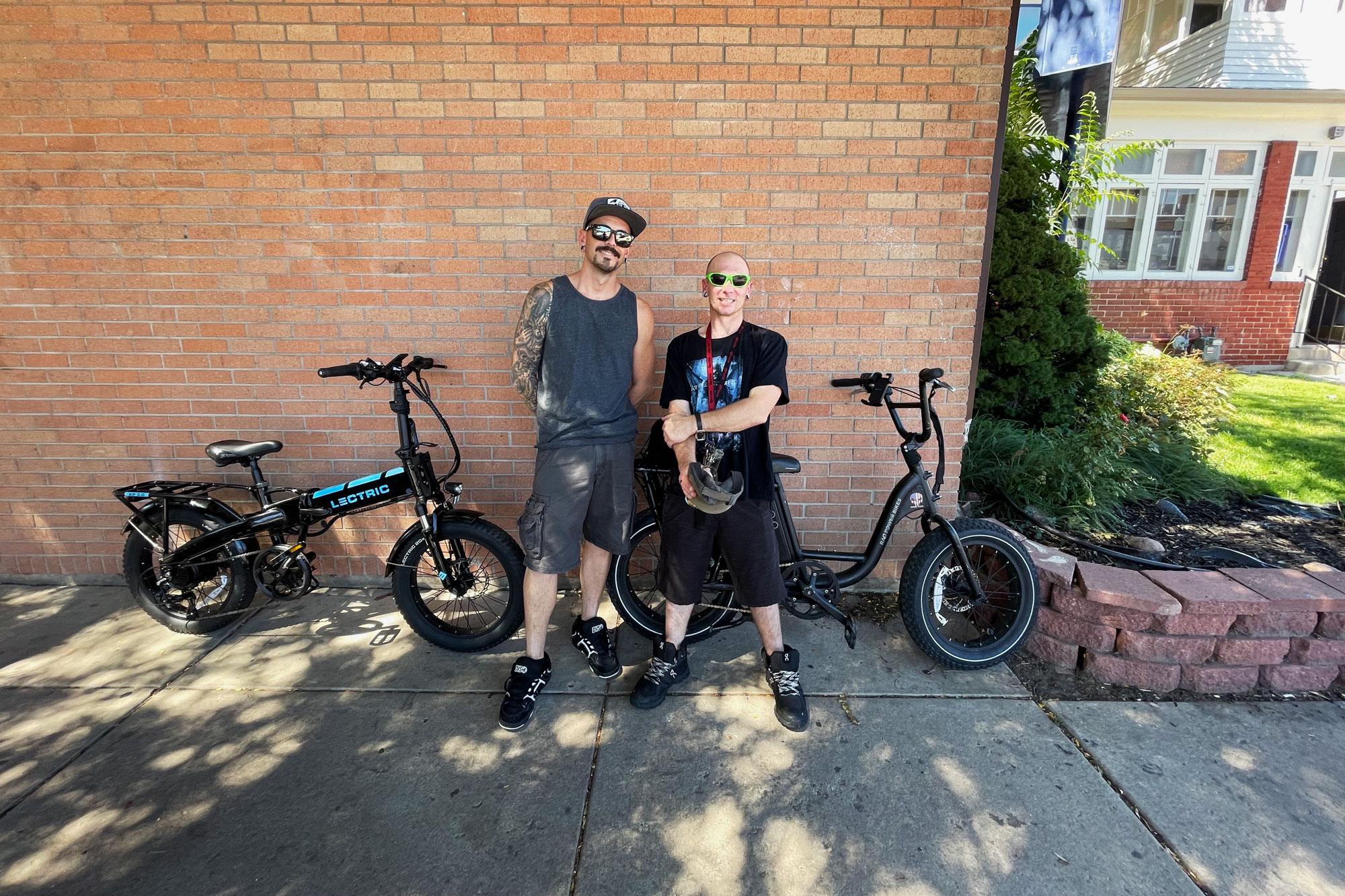 Two men stand with their e-bikes in front of a brick wall.