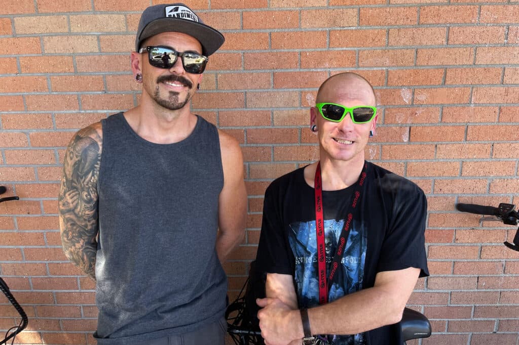 Two men stand against a brick wall.