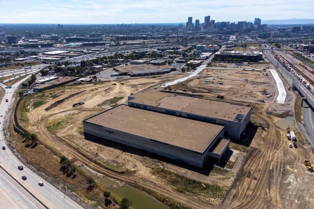 An aerial view of a construction site, covered in dirt and populated only by a few grey concrete buildings. Denver's skyline rises in the background.