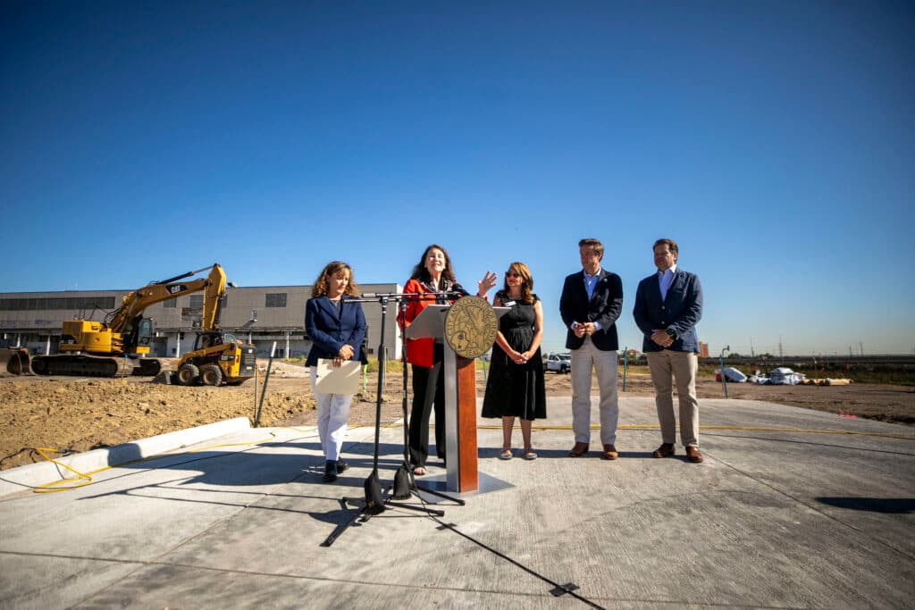 A woman in a red jacket speaks at a microphone, surrounded by three other people dressed like they're important. There's construction equipment behind them.