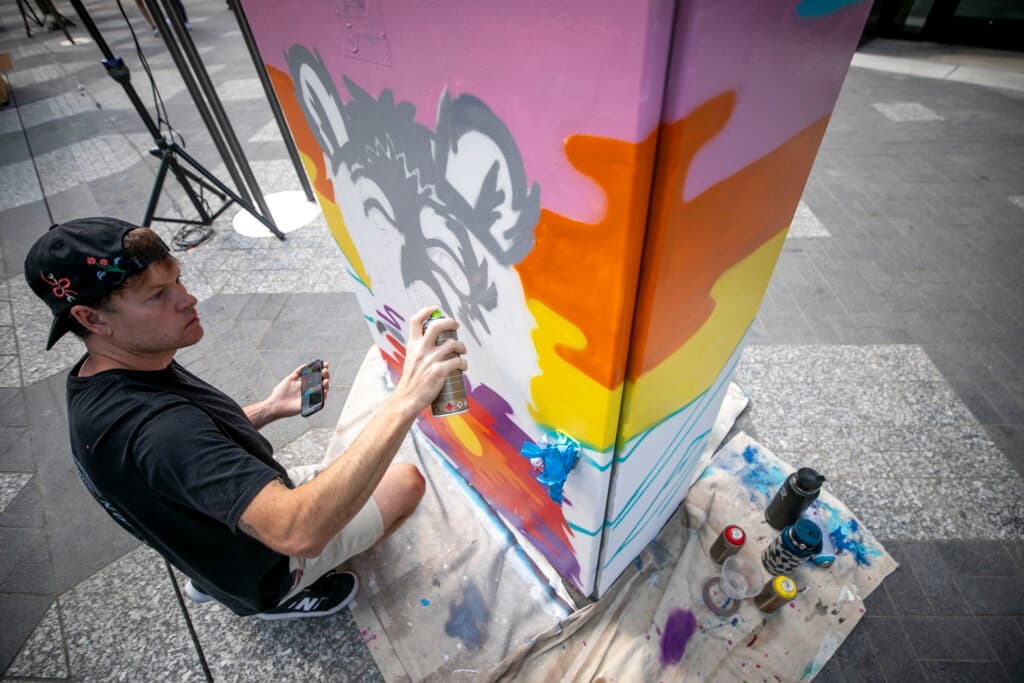 A man in a ball cap kneels in front of a colorfully painted utility box, pointing a spray paint can at what appears to be a wolf coming together on the front. He's surrounded by paint cans.