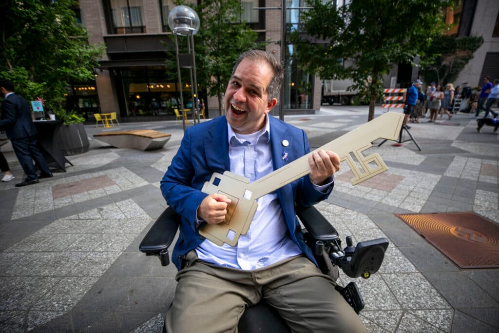 A man in a blue suit jacket holds a giant golden key, playing it like a guitar.