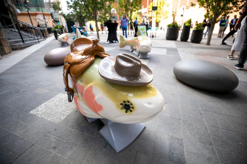 A shiny pink, white and beige sculpture of trout. It's wearing a cowboy hat and a saddle; other fish &quot;swim&quot; along the brick ground behind it.