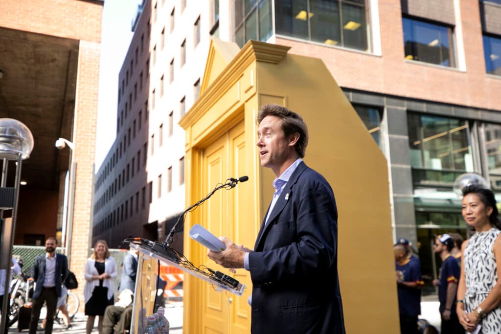 A man in a suit jacket smiles as he speaks at a podium. Office buildings and a freestanding golden doorway are behind him.