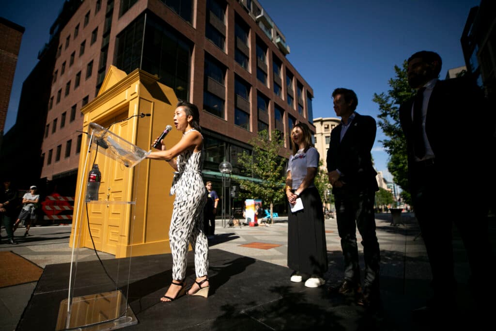 A woman in a black and white jumpsuit speaks at a microphone, surrounded by people silhouetted in the shade. A golden doorway and office buildings are behind her.