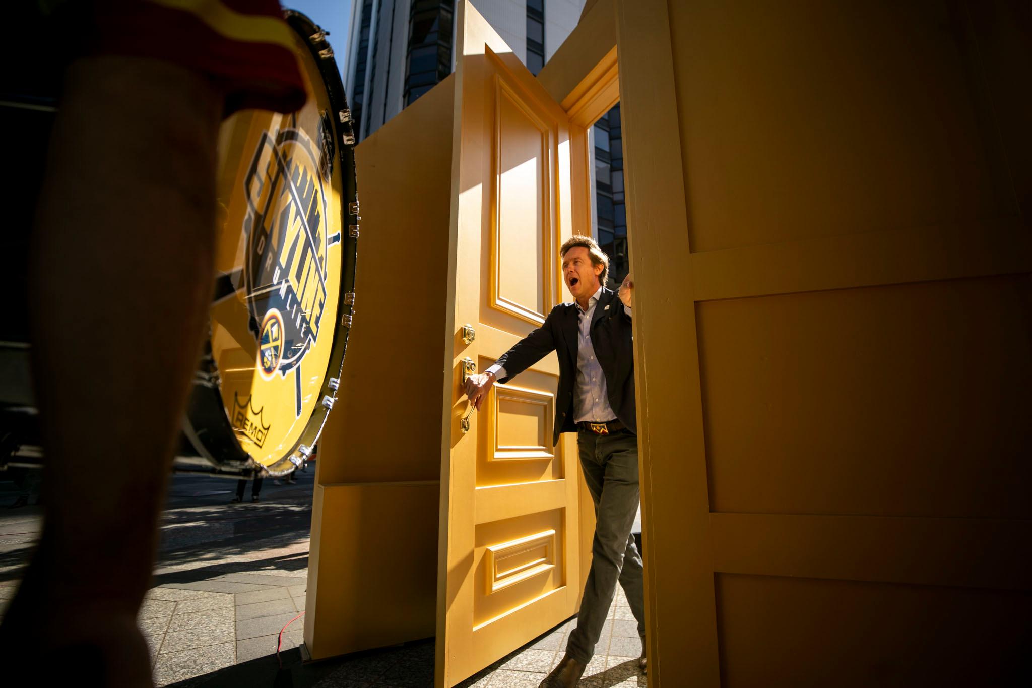 A man in a suit jacket walks through a golden doorway, backlit by the morning sun. His mouth is agape in what could be excitement.