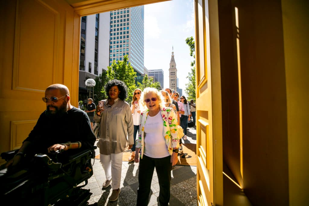 A line of people, backlit by the morning sun, pass through a gateway of gold. A clocktower and office buildings rise in the distance.