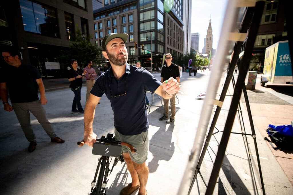 A man holds a bike as he reaches forward with a little green sticker in his hand, about to stick it to a board on an easel in the foreground. The downtown clocktower rises in the distance.