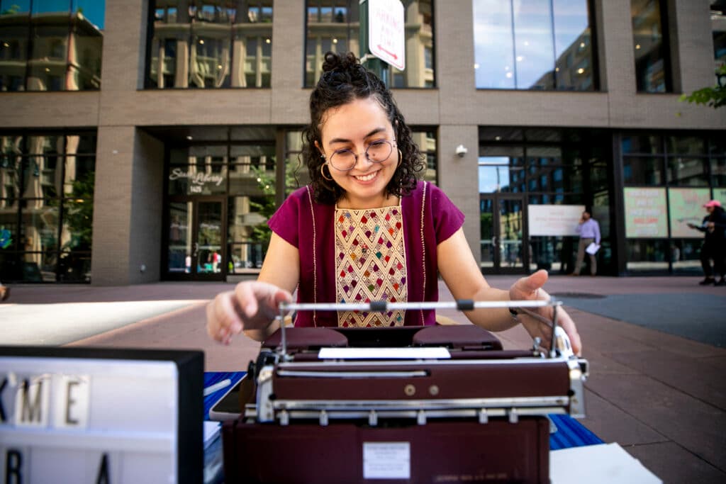 A woman in a crimson shirt smiles as she reaches forward to adjust a typewriter.