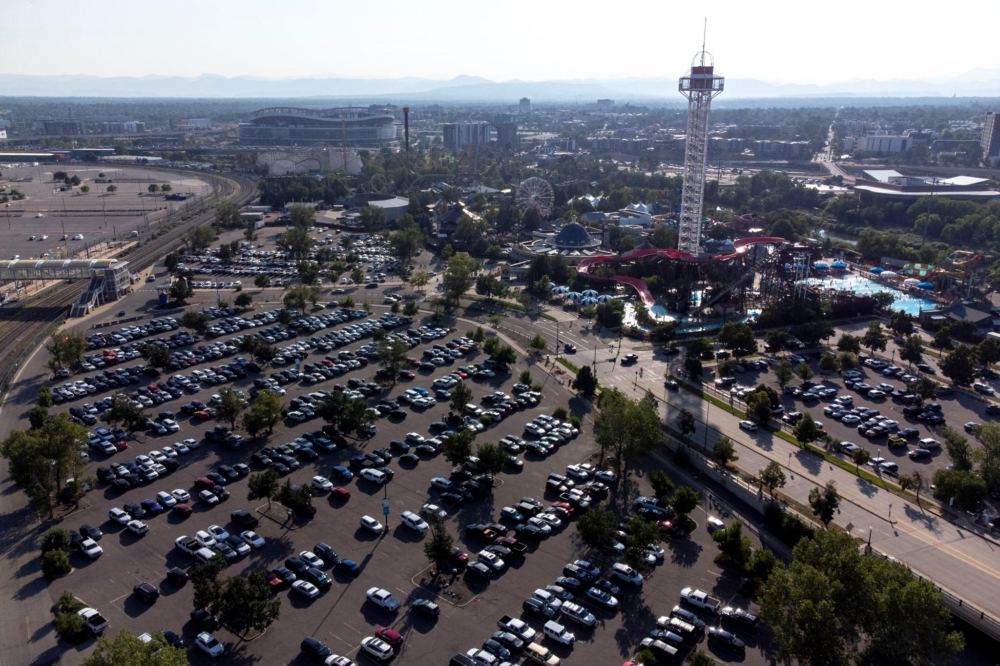An aerial view of Elitch Gardens, its white view tower breaking the mountain-lined horizon, and a big, full parking lot.