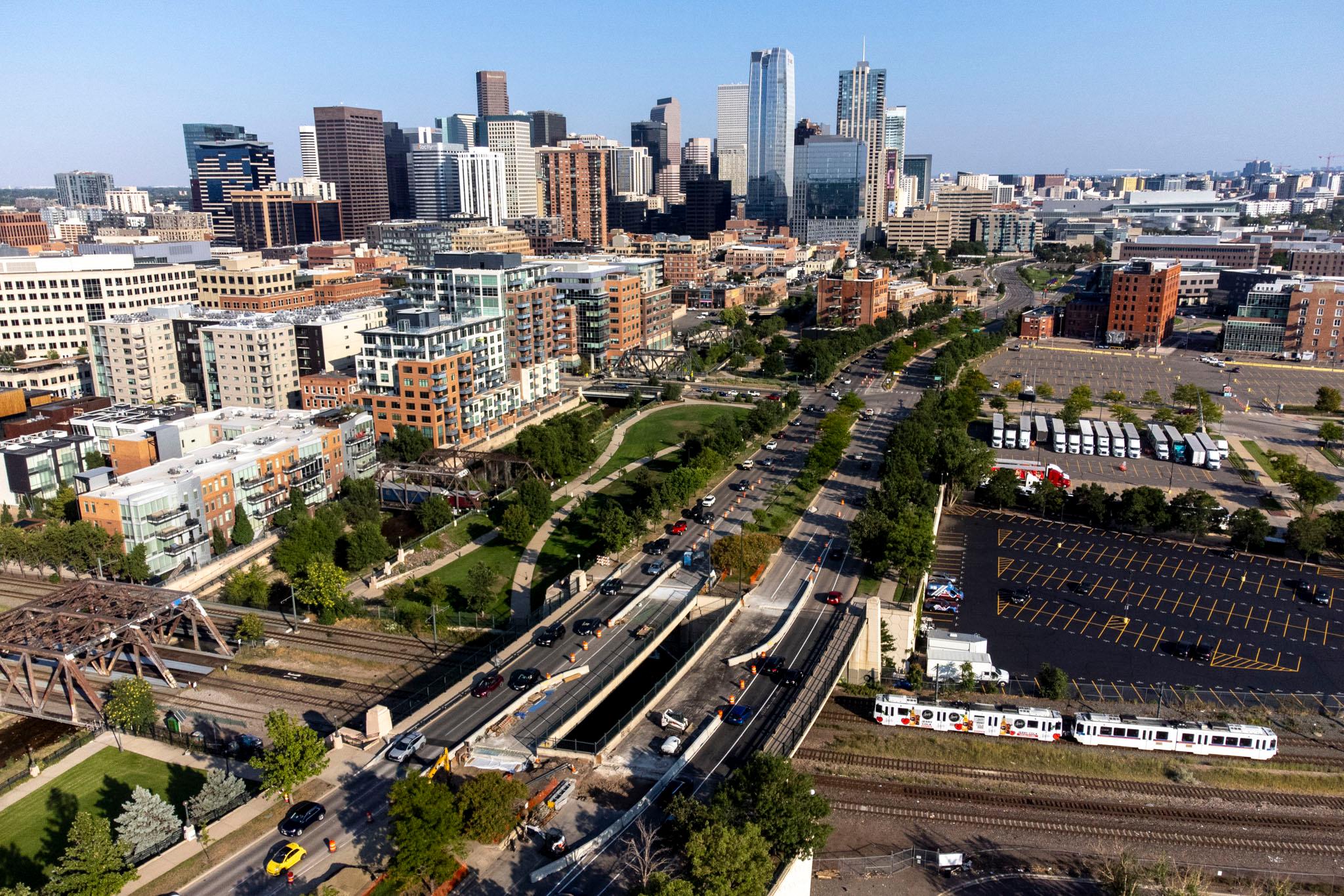 An aerial view of downtown Denver, with Speer Boulevard's tree-lined asphalt running through the center of the frame into the distance.