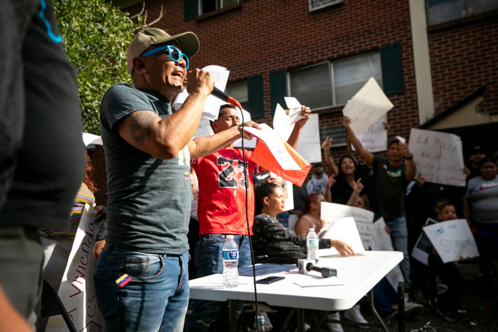 A man in a ball cap and shades yells into a microphone, as people holding signs around him raise their arms and shout, too.