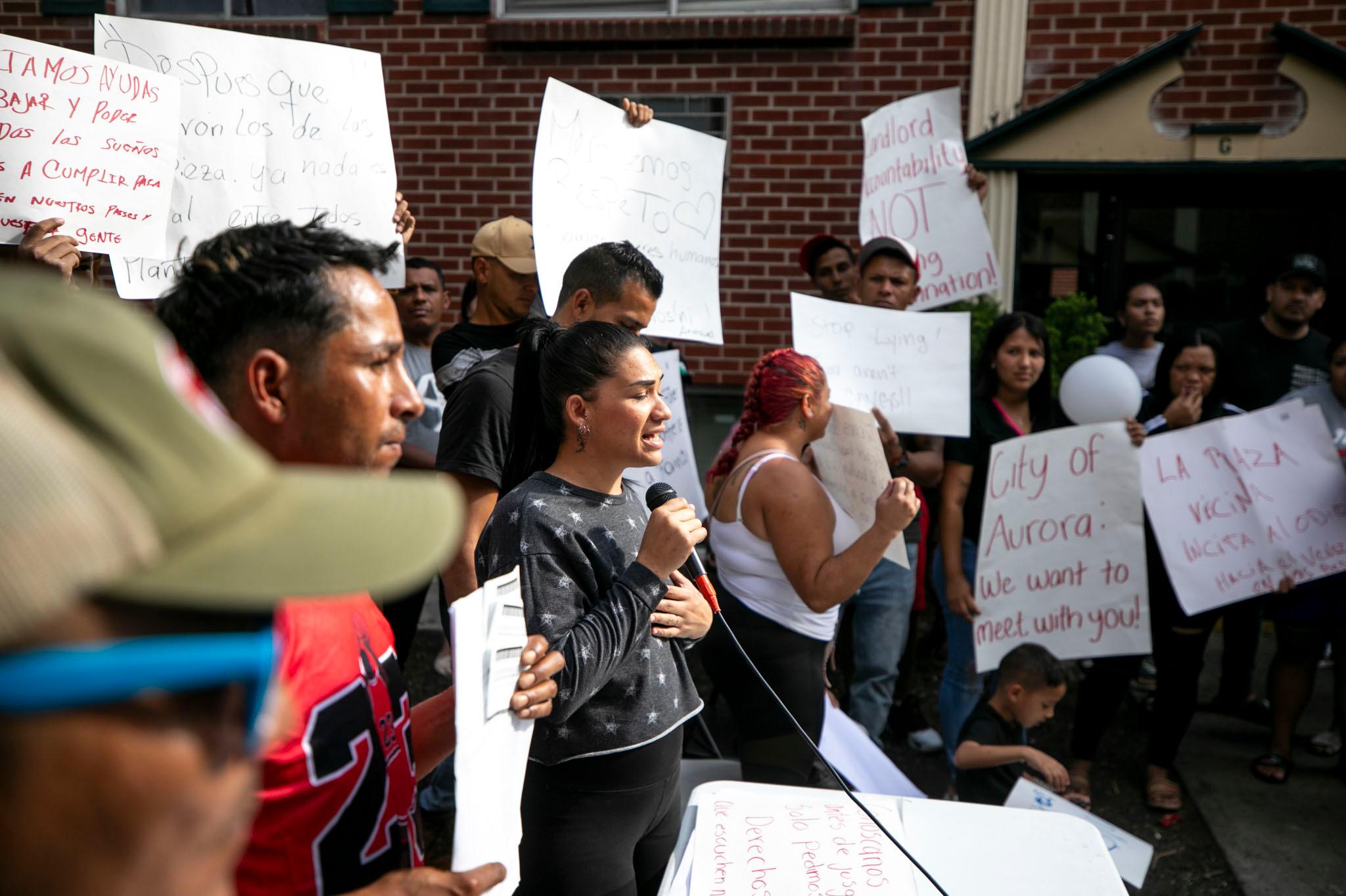 A woman in a grey sweater looks pained as she speaks into a microphone. People around her hold up signs with handwritten notes on them.