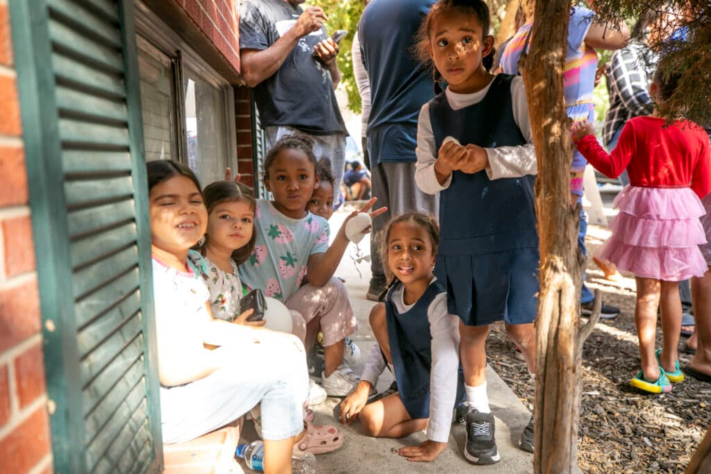 Six little girls, two in matching navy dresses, smile towards a camera as they sit on a windowsill under a bush.